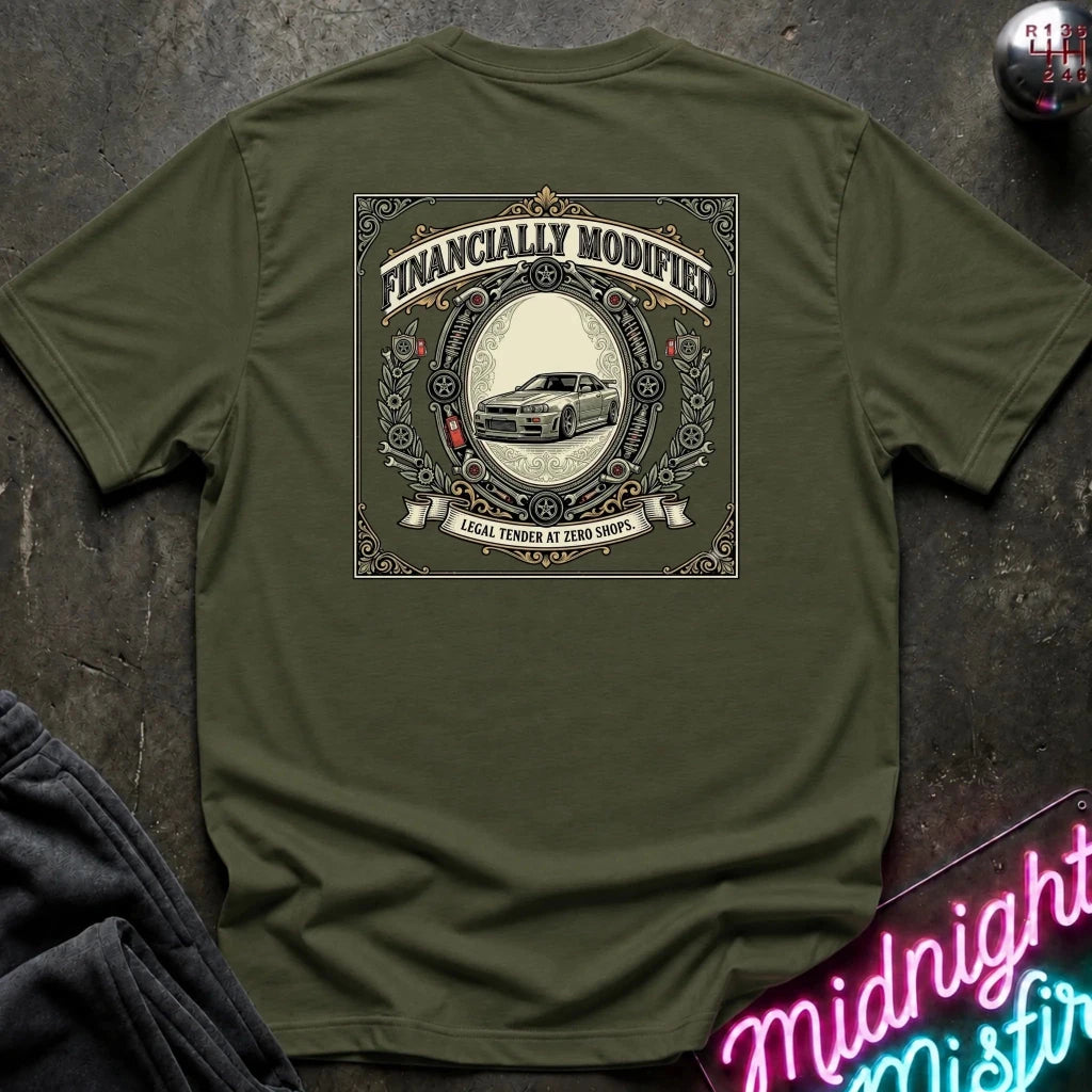 An olive green short-sleeve t-shirt laid flat on a dark textured surface, featuring a large vintage-style graphic on the back with the text 'FINANCALLY MODIFIED' and a car illustration. A neon sign and folded dark fabric are partially visible in the corners.