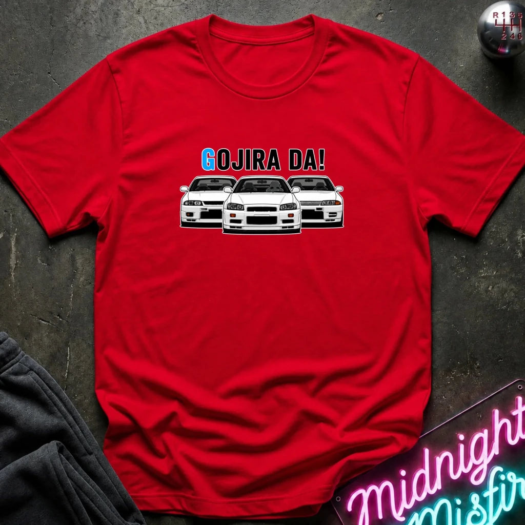 A red short-sleeve crewneck t-shirt featuring a graphic of three white Nissan Sentra cars with the text 'GOJIRA DA!' printed above them. The shirt is displayed flat on a dark textured surface with a gray sweatshirt corner and a neon sign in the background.