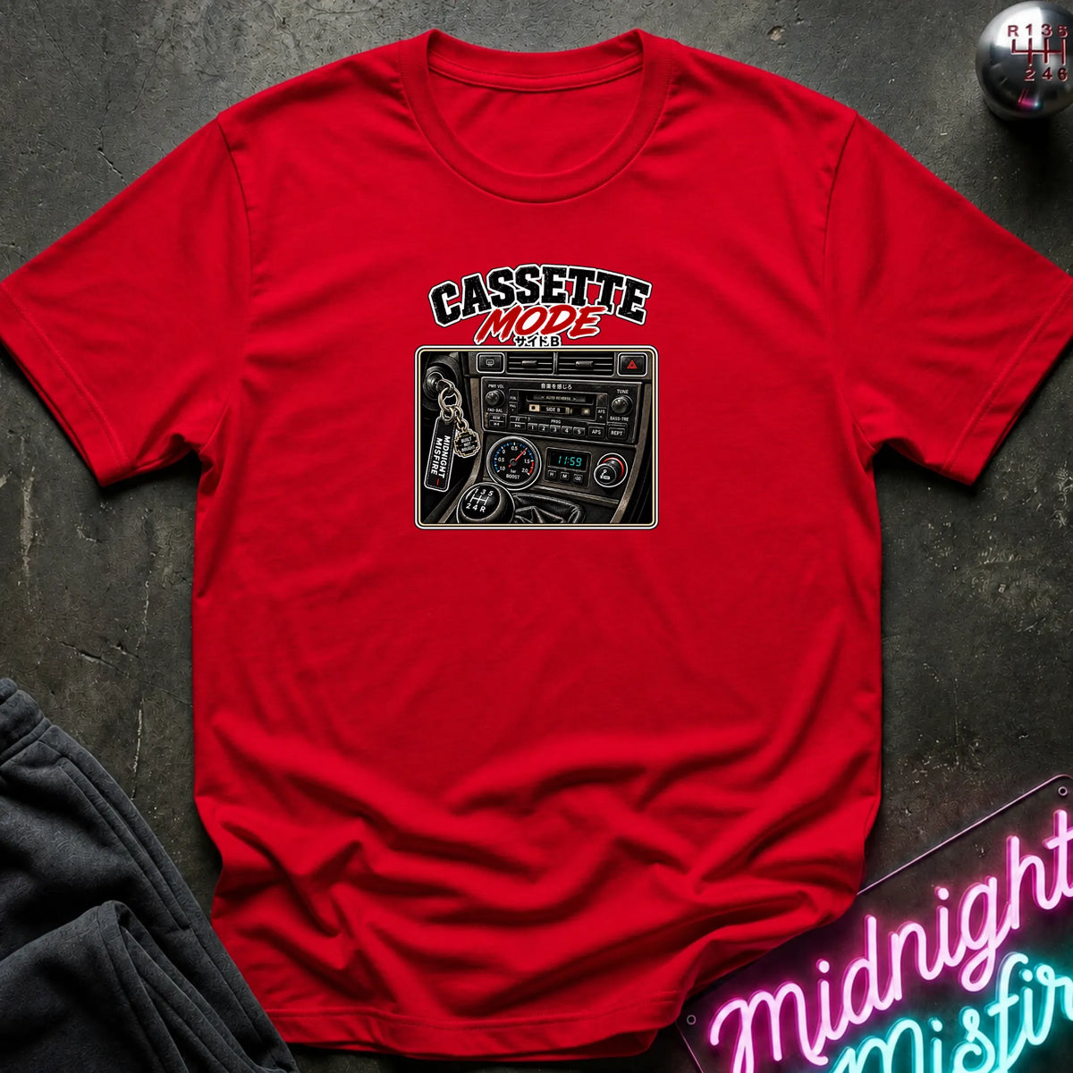 A red short-sleeve t-shirt laid flat on a dark textured surface, featuring a graphic print of a cassette radio with the text 'CASSETTE MODE' in black and red. A neon sign and dark pants are partially visible as props.