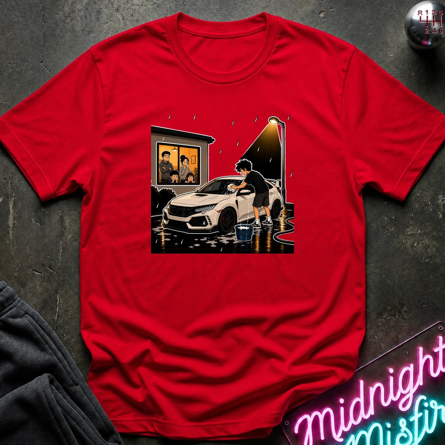 A red short-sleeve crewneck t-shirt with a graphic print of a person washing a white sports car in the rain, styled flat on a dark textured background with a neon sign and dark sweatpants partially visible.
