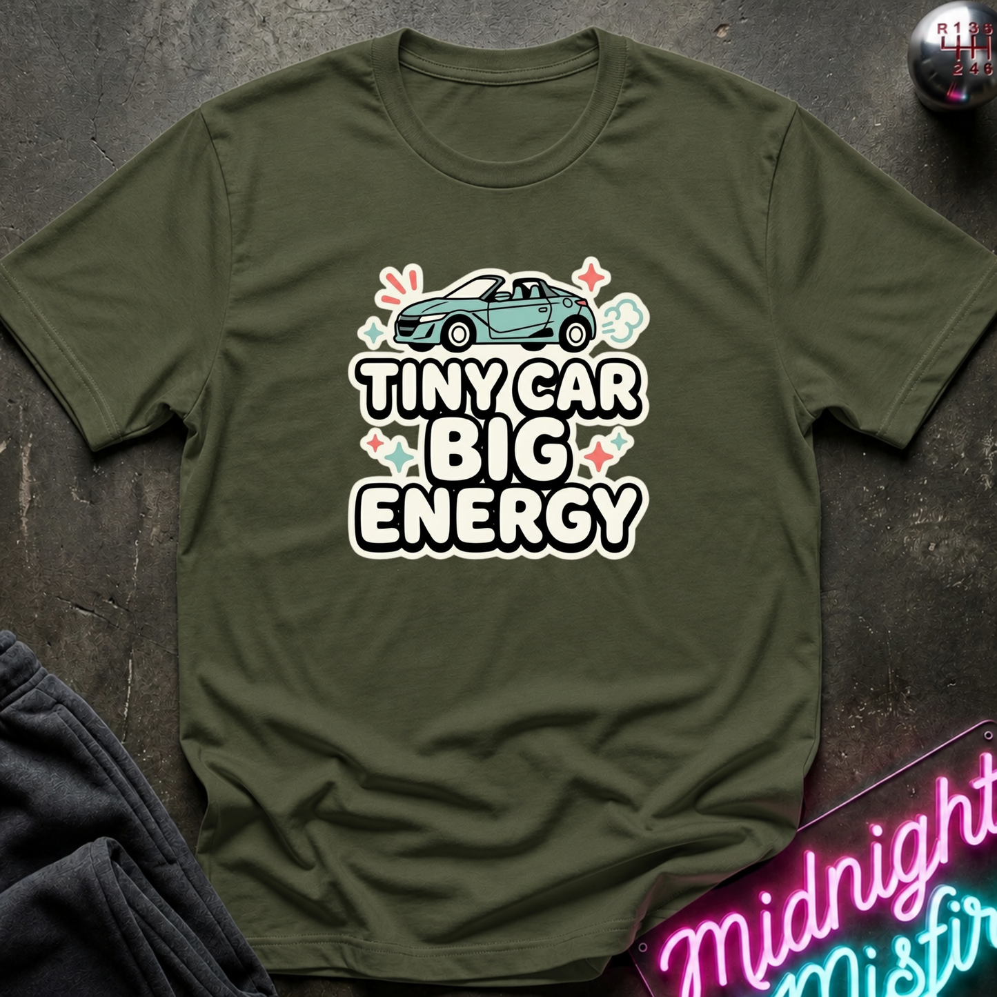 An olive green short-sleeve t-shirt with a graphic print of a small teal convertible car and the text 'TINY CAR BIG ENERGY' in white with a black outline. The shirt is laid flat on a dark textured surface with a neon sign and dark fabric partially visible in the corners.