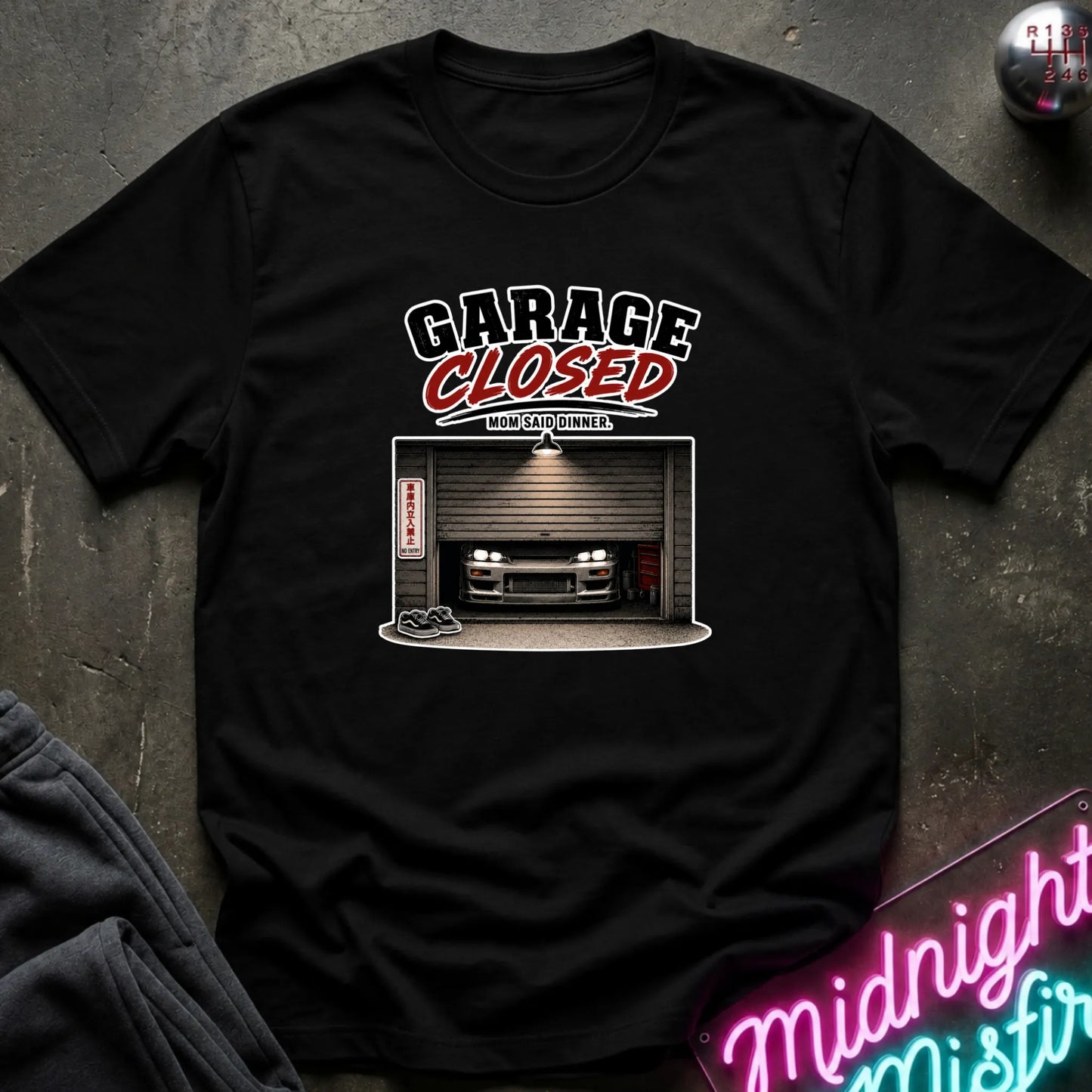 A black short-sleeve graphic t-shirt laid flat on a dark textured surface. The graphic features the words 'GARAGE CLOSED' in white and red, above an illustration of a car parked inside a closed garage.