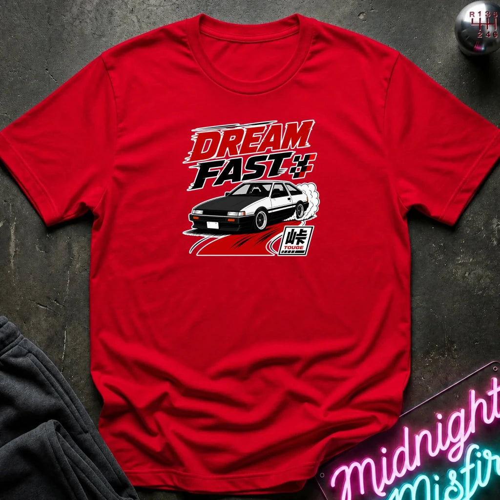 A red short-sleeve graphic T-shirt laid flat on a dark textured surface. The shirt features a red, black, and white 'DREAM FAST' graphic with a car illustration.