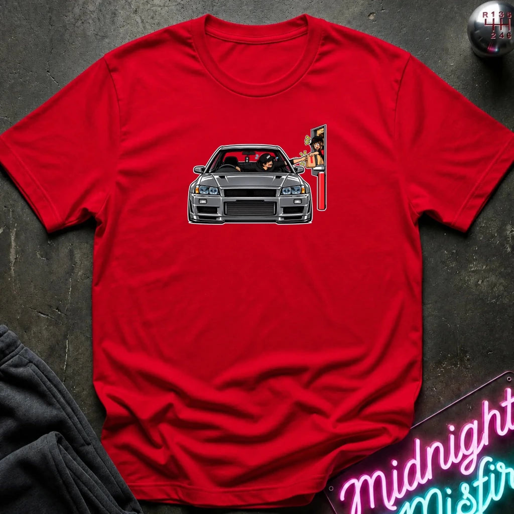 A red short-sleeve crewneck t-shirt featuring a graphic of a gray sports car with a driver and a street sign character, laid flat on a dark textured surface with lifestyle props.