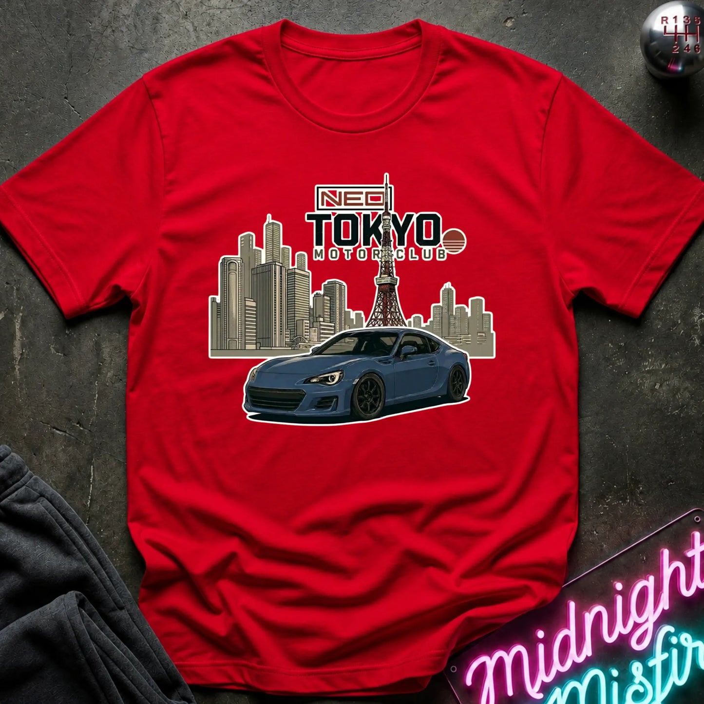 A red short-sleeve graphic T-shirt laid flat on a dark textured surface. The shirt features a printed design of a blue sports car in front of a Tokyo city skyline with the Tokyo Tower, and text reading 'NEO TOKYO MOTORCLUB'.