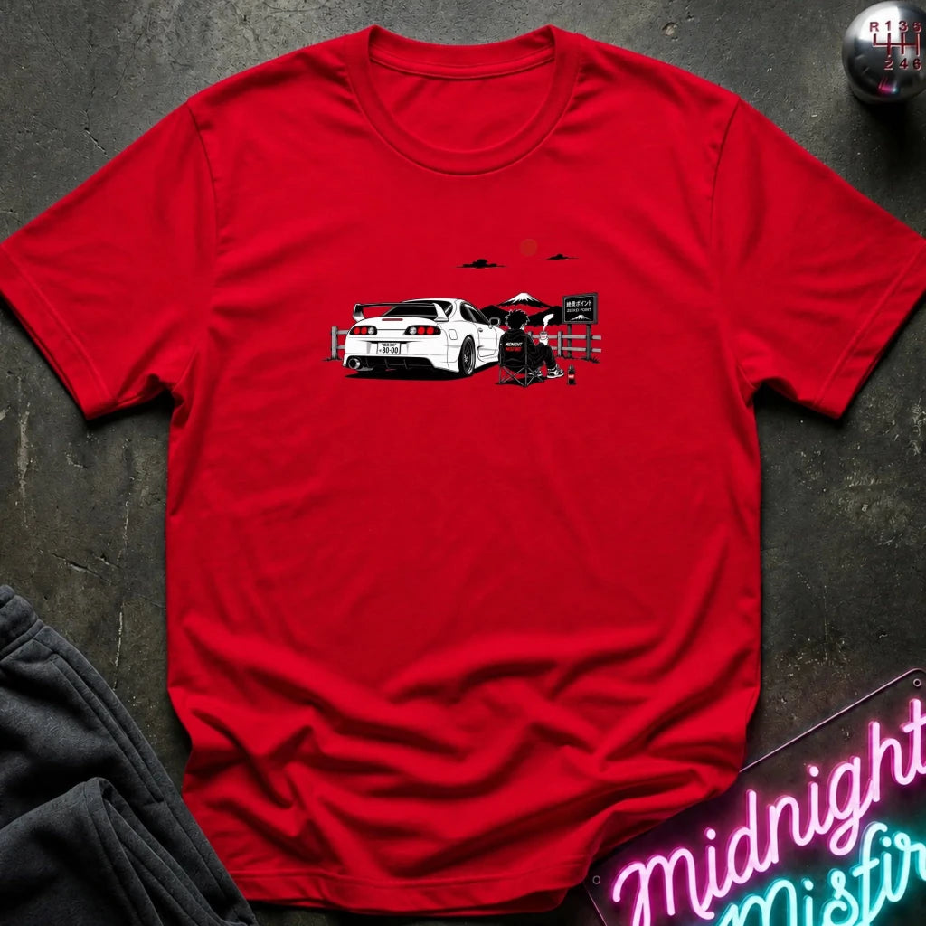 A red short-sleeve graphic T-shirt laid flat on a dark textured surface. The shirt features a printed illustration of a white sports car with a red sun and mountain graphic above it.