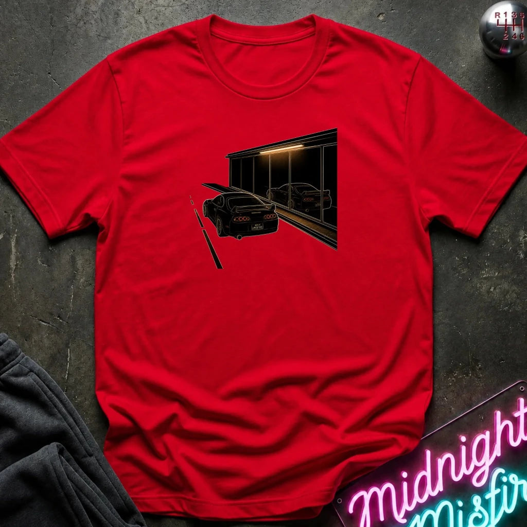 A red short-sleeve t-shirt laid flat on a dark textured surface, featuring a graphic print of a black sports car in front of a dark building with warm light. 
