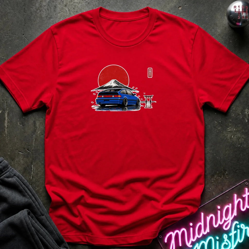 A red short-sleeve crewneck t-shirt laid flat on a dark textured surface. The center of the shirt features a graphic print of a blue sports car in front of a red sun and mountain, with a small red torii gate and a vertical red character mark.