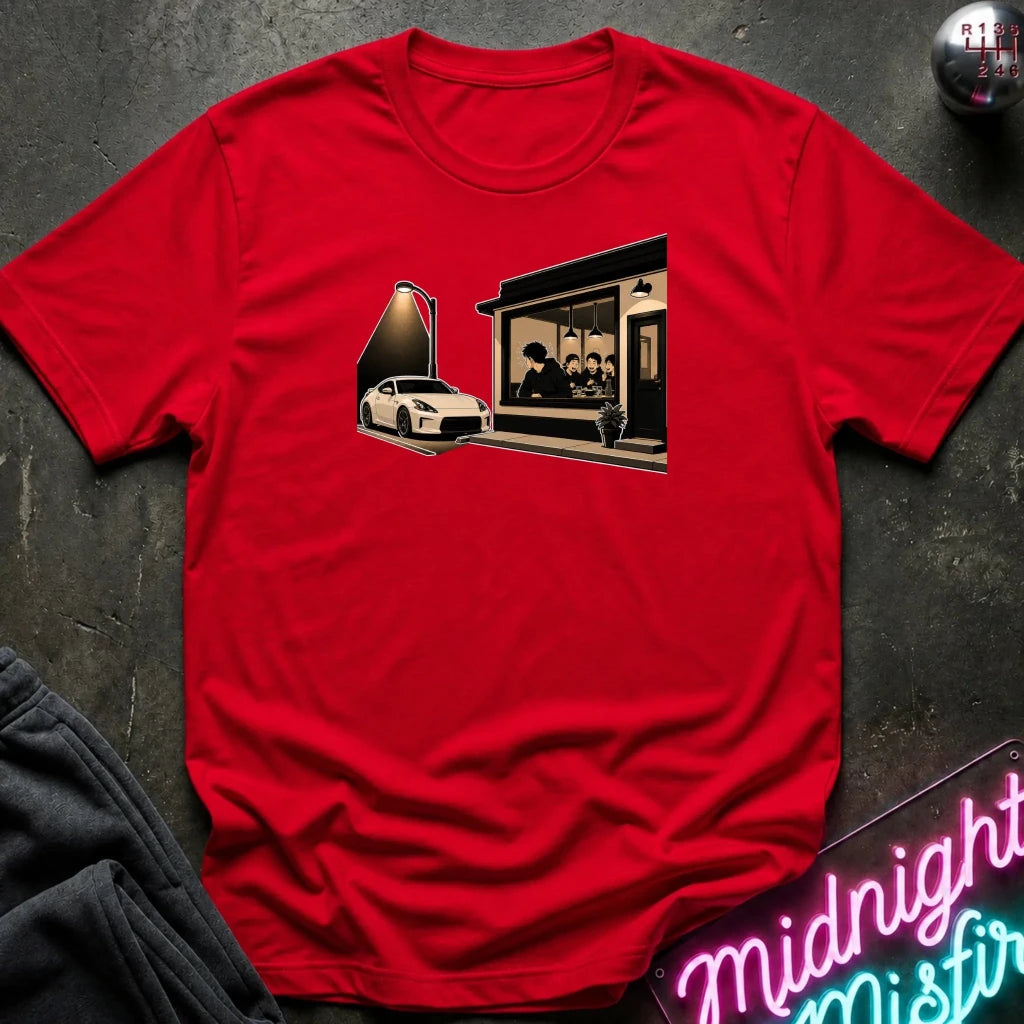 A red short-sleeve crewneck t-shirt laid flat on a dark textured surface, featuring a graphic print of a white sports car parked in front of a dark building. 