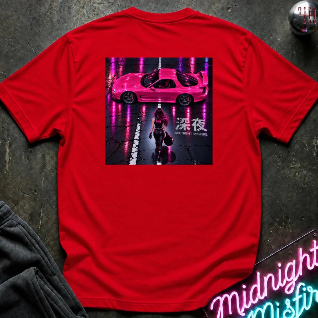 A bright red short-sleeve t-shirt laid flat on a dark textured surface, featuring a large rectangular graphic print on the back with a pink sports car, a person, and neon-style text. Partially visible props include dark gray pants in the lower left and a neon sign in the lower right.