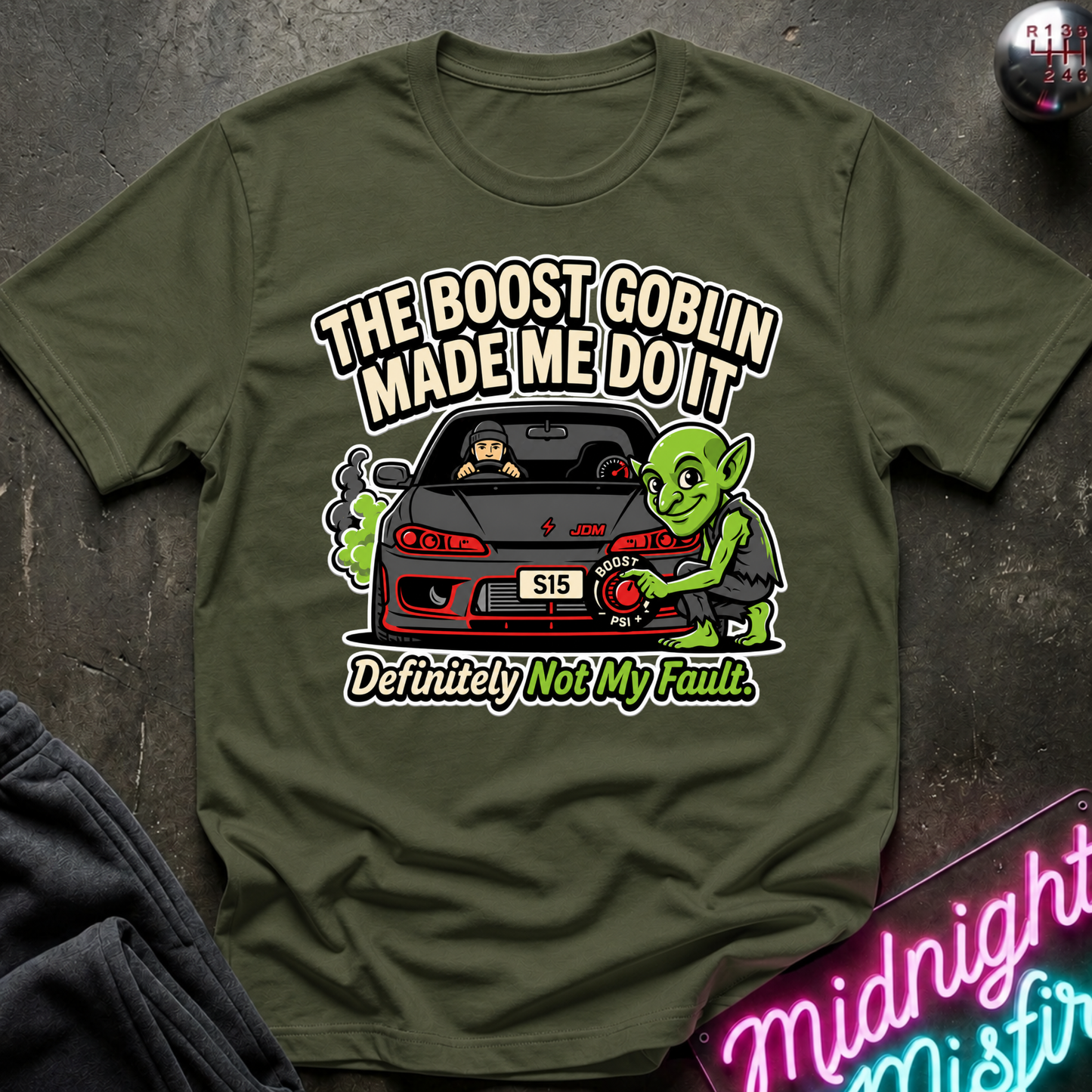An olive green graphic t-shirt laid flat on a dark textured surface. The graphic features a green character next to a black sports car with the text 'THE BOOST GOBLIN MADE ME DO IT' above and 'Definitely Not My Fault' below.