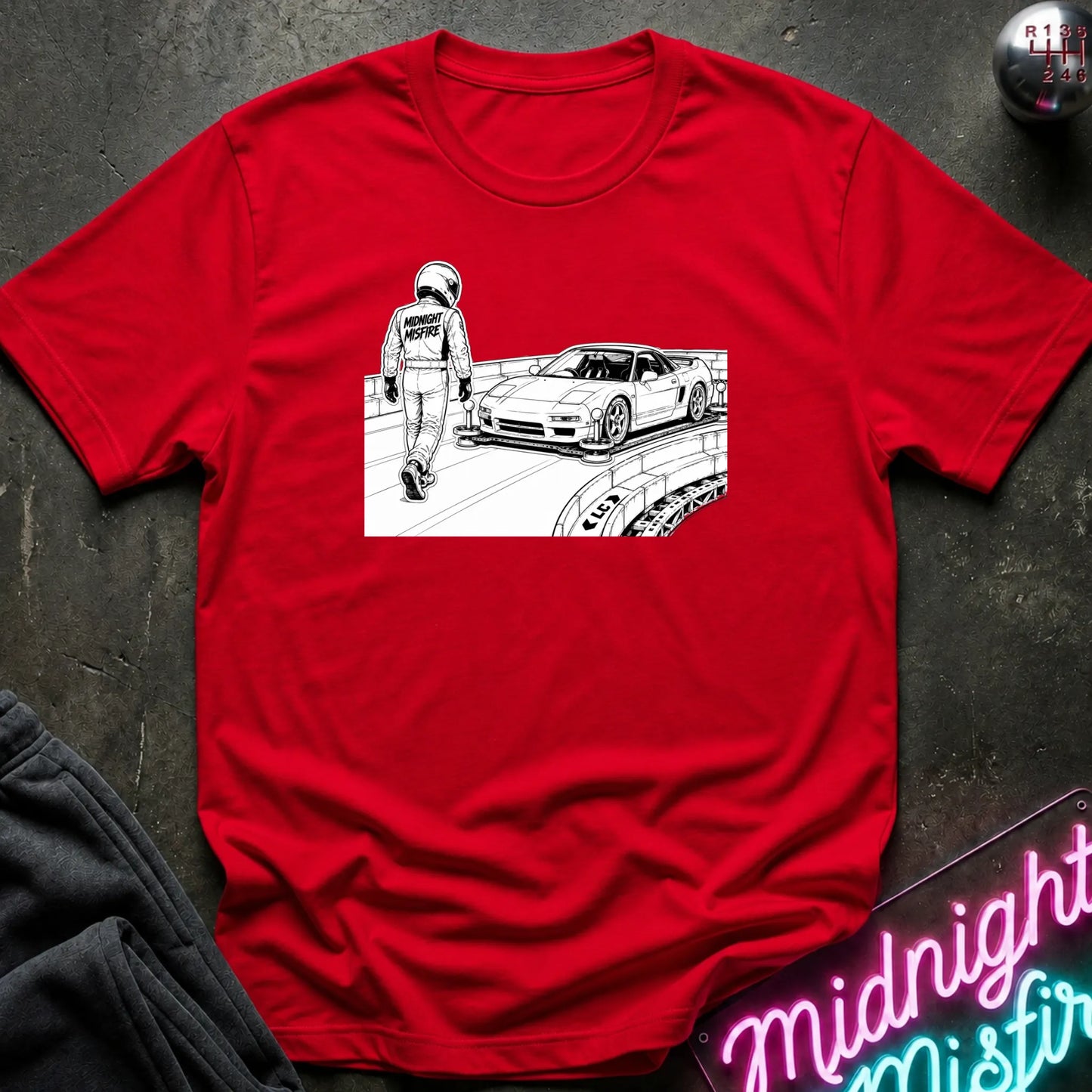 A red short-sleeve crewneck t-shirt laid flat on a dark textured surface. The shirt features a black and white graphic print on the chest depicting a race car and a driver walking.