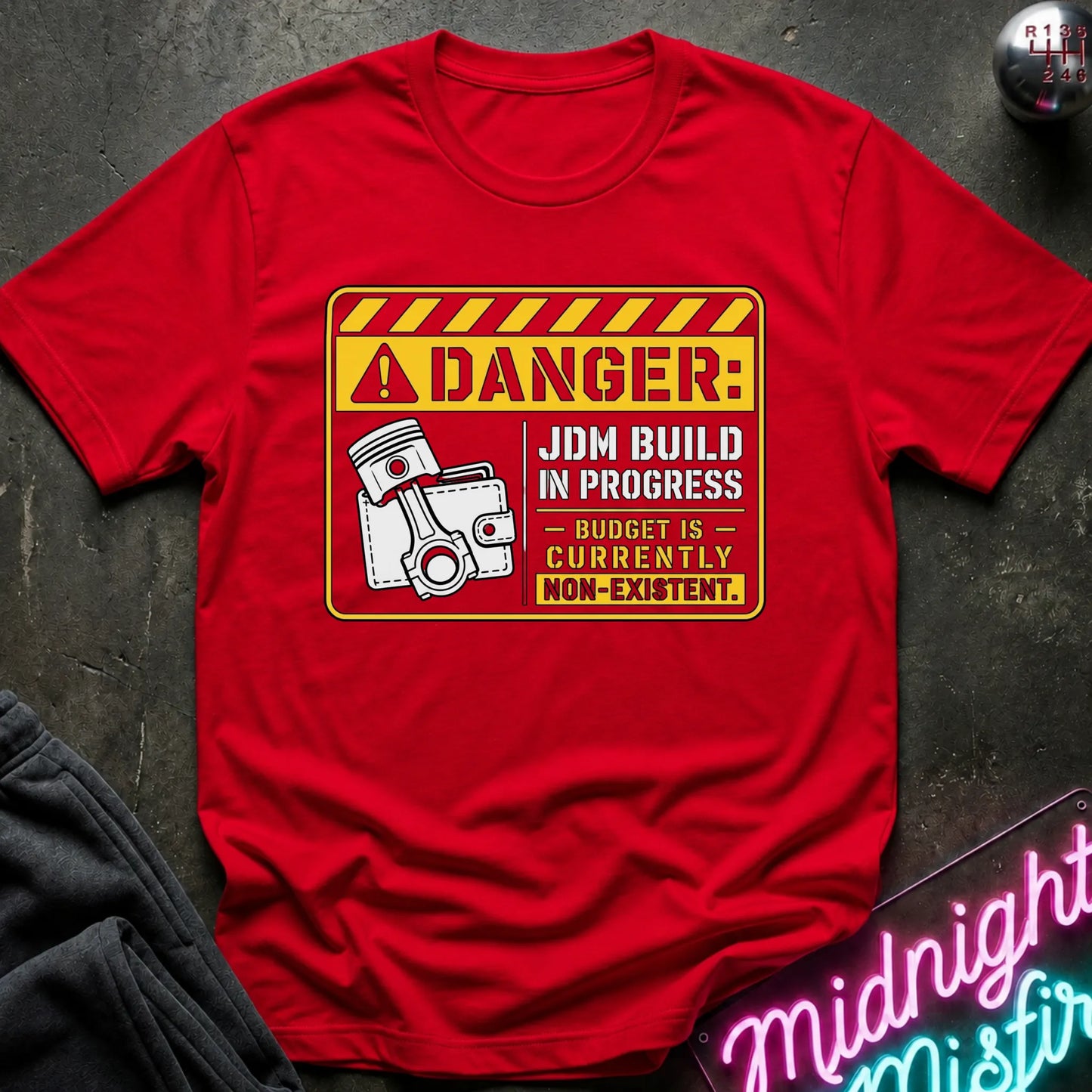 A red short-sleeve graphic T-shirt laid flat on a dark textured surface. The shirt features a yellow and white warning-sign style print with the text 'DANGER: JDM BUILD IN PROGRESS - BUDGET IS - CURRENTLY NON-EXISTENT'.