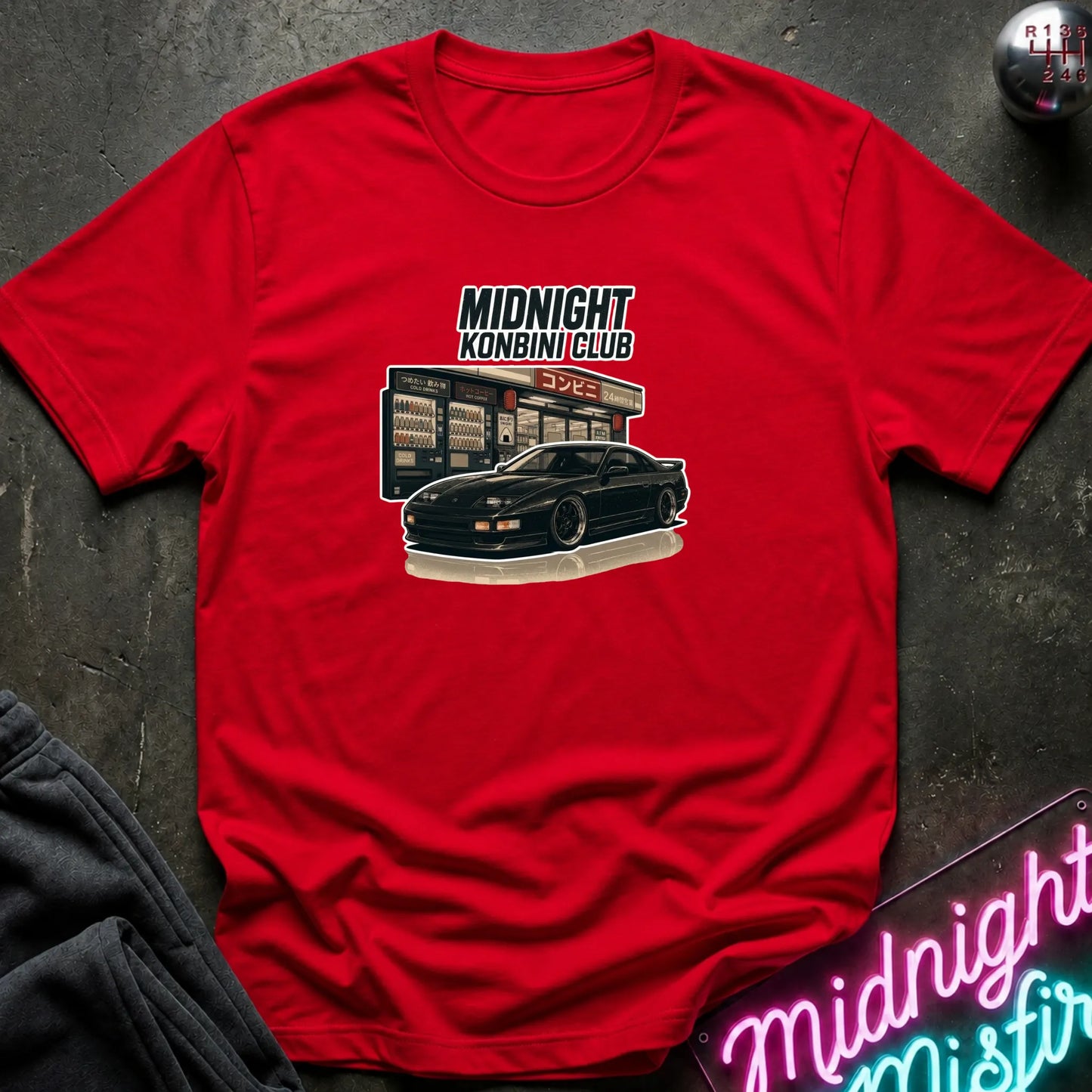 A red short-sleeve graphic t-shirt laid flat on a dark textured surface. The graphic features the text 'MIDNIGHT KONBINI CLUB' above an illustration of a black sports car parked in front of a convenience store.