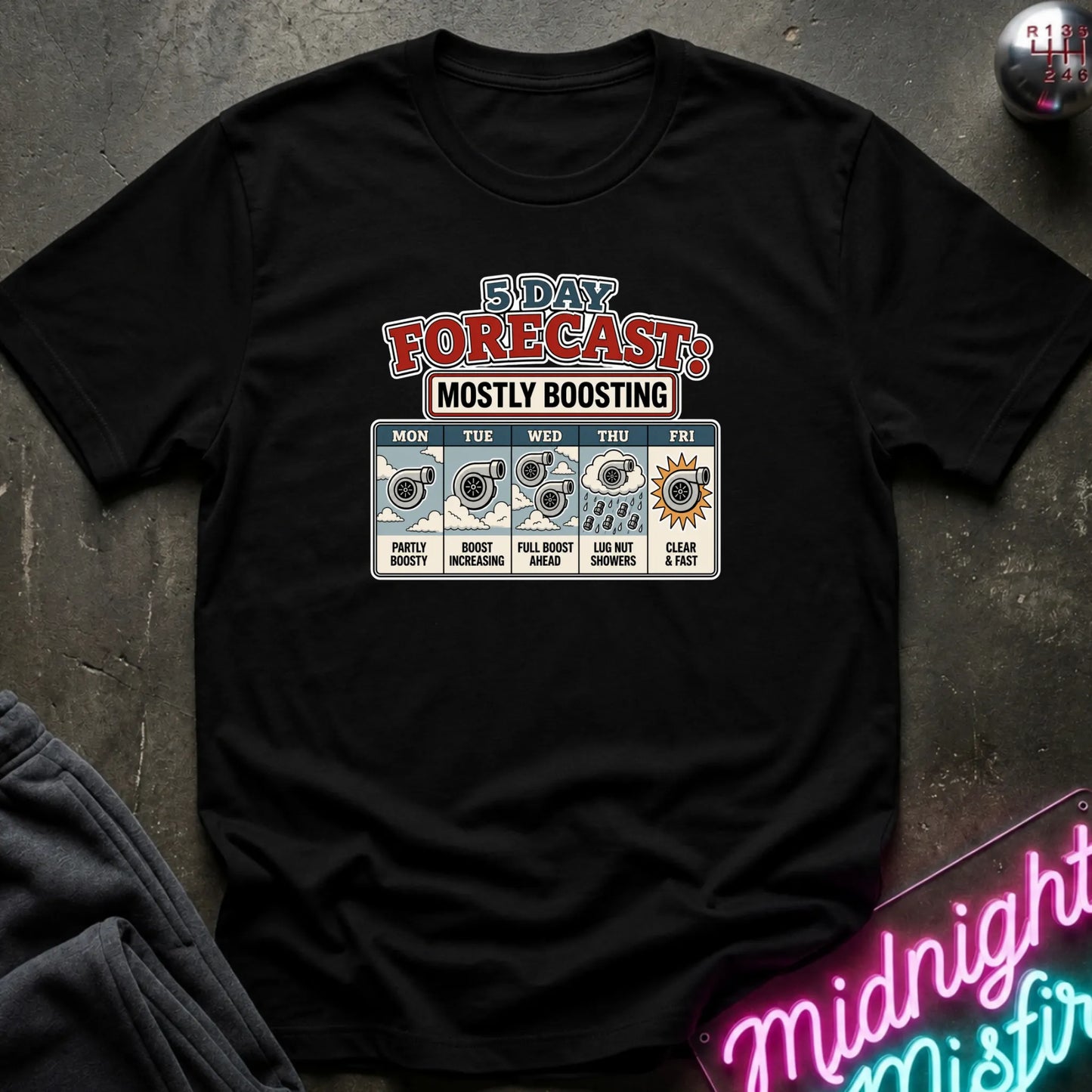A black short-sleeve graphic t-shirt laid flat on a dark textured surface. The shirt features a '5 Day Forecast: Mostly Boosting' graphic with daily weather icons and text from Monday to Friday.