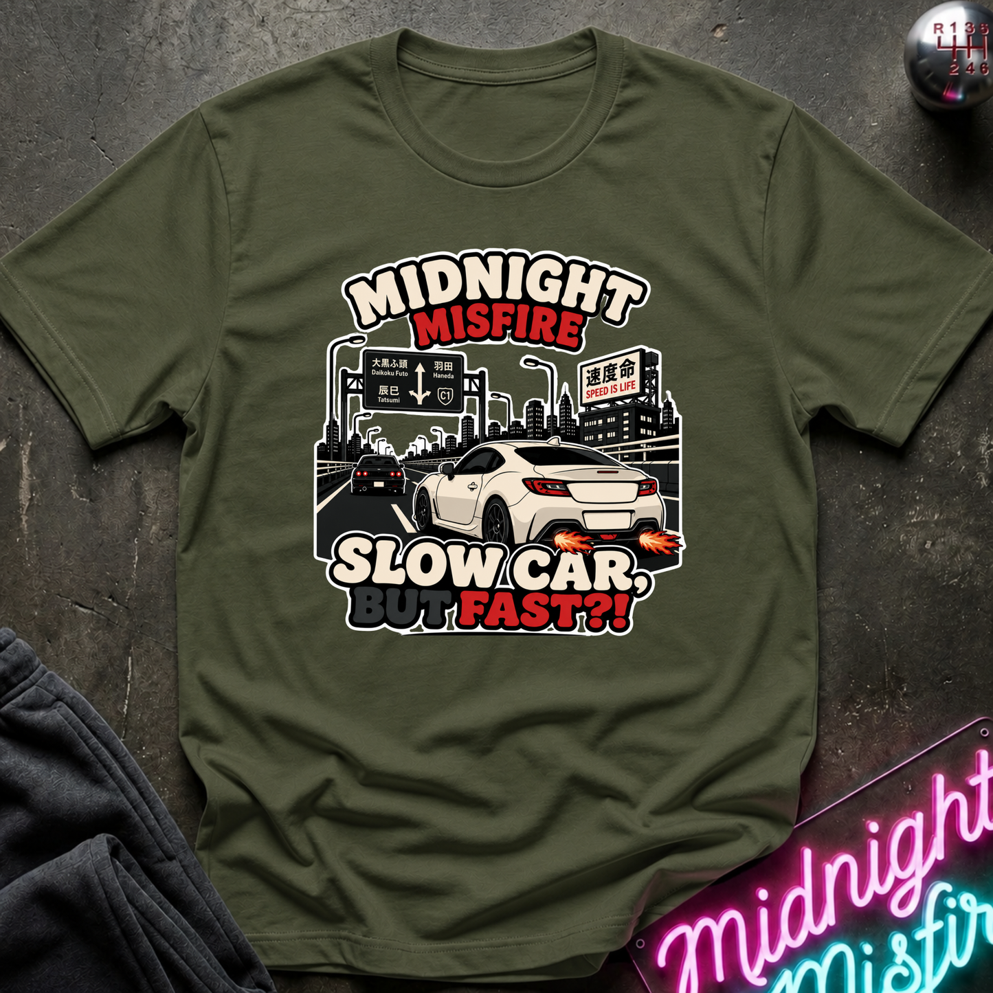 An olive green graphic t-shirt laid flat on a dark textured surface. The graphic features a white sports car and city street scene with the text 'MIDNIGHT MISFIRE SLOW CAR, FAST?!!' in white, red, and black.