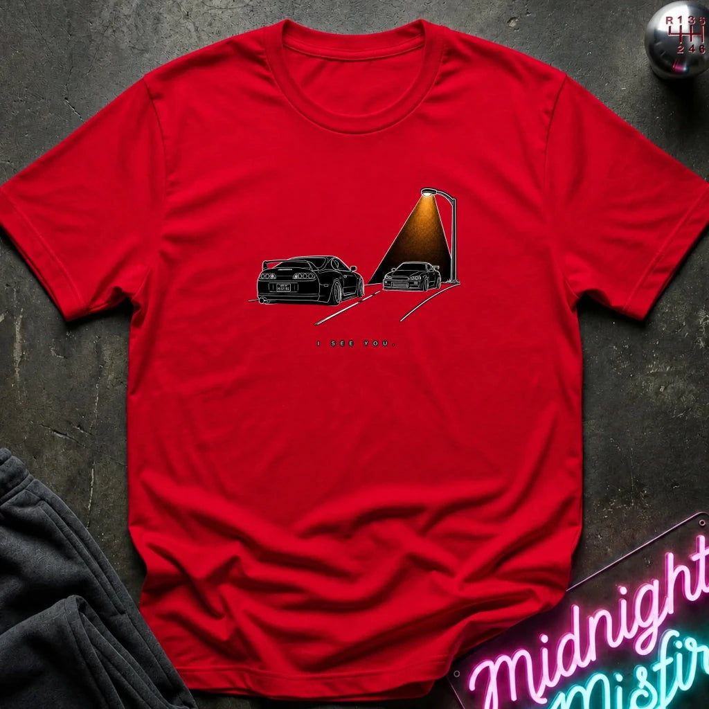 A red short-sleeve t-shirt laid flat on a dark textured surface, featuring a graphic print of two black sports cars passing each other.