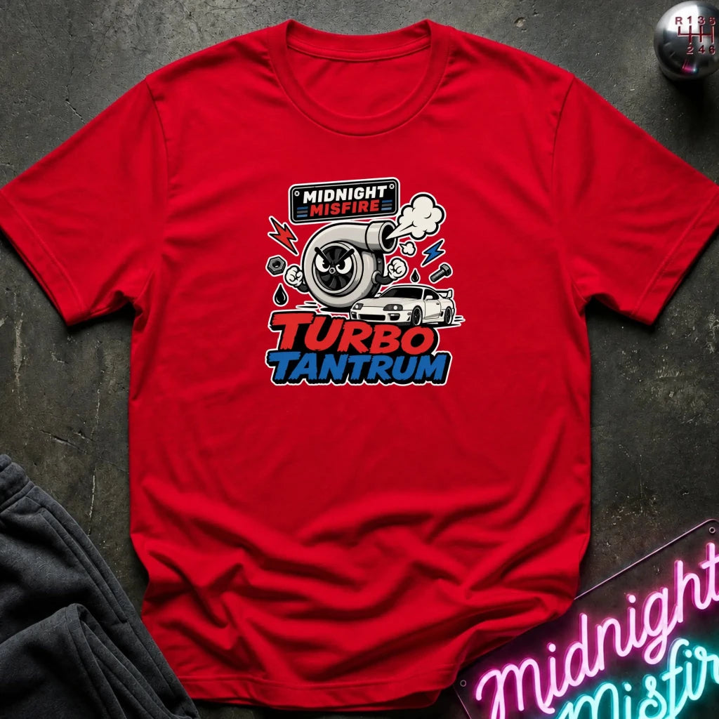 A red short-sleeve graphic T-shirt laid flat on a dark textured surface. The shirt features a central graphic with a black-and-white car, a large silver turbocharger, smoke, and the text 'MIDNIGHT MISFIRE' and 'TURBO TANTRUM' in red, white, and blue.