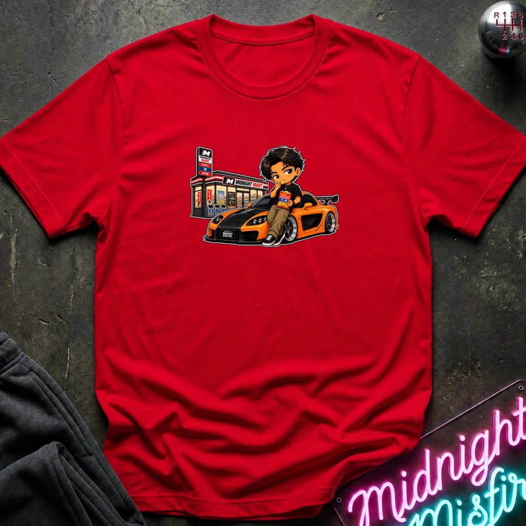 A red short-sleeve crewneck t-shirt laid flat on a dark textured surface, featuring a cartoon graphic of a man sitting on a orange and black sports car next to a store.