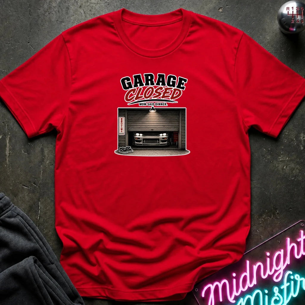 A red  short-sleeve graphic t-shirt laid flat on a dark textured surface. The graphic features the text 'GARAGE CLOSED' in black and red, above an illustration of a car parked inside a garage.