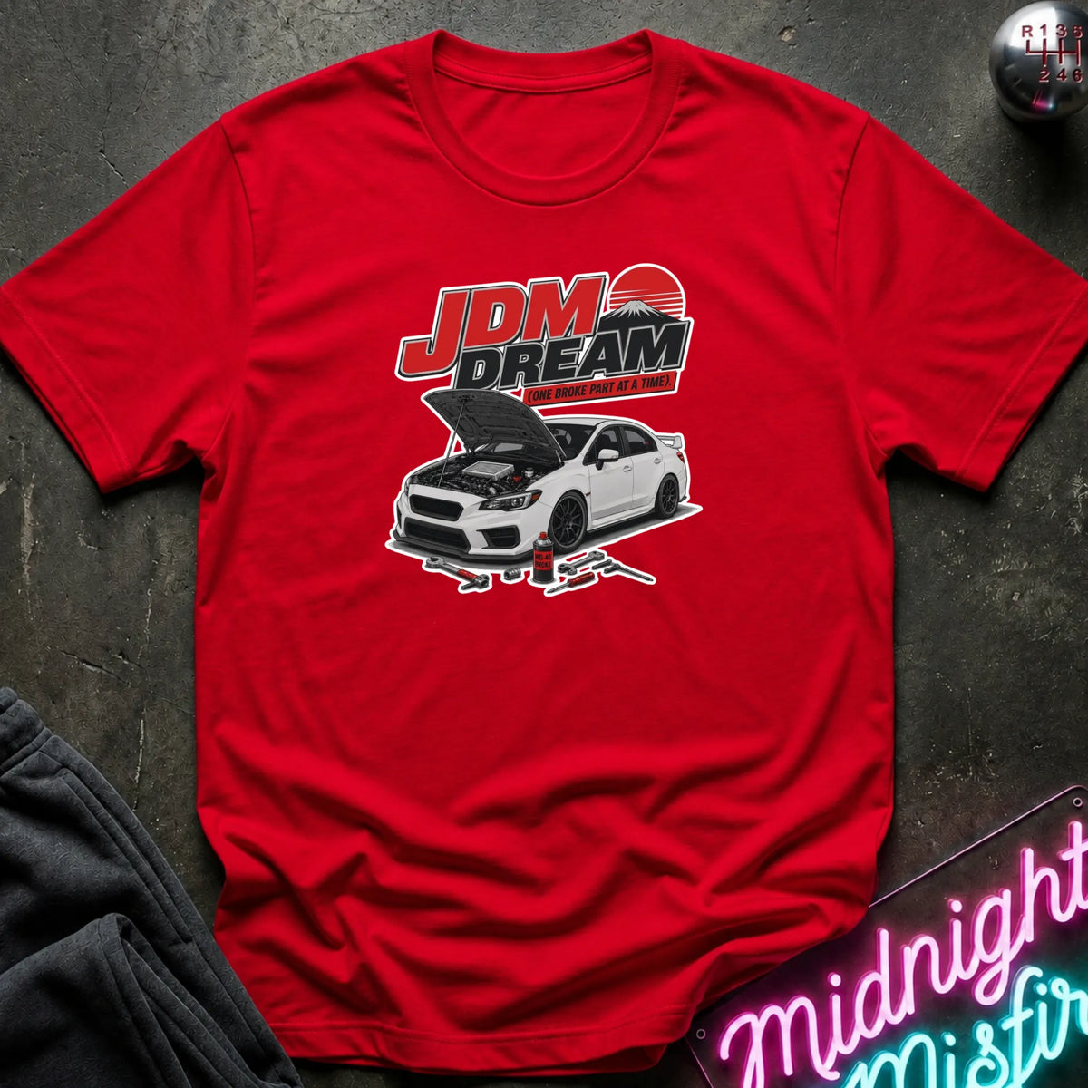A red short-sleeve graphic T-shirt laid flat on a dark textured surface. The shirt features a white car with its hood open beneath the text 'JDM DREAM' in red, white, and black.