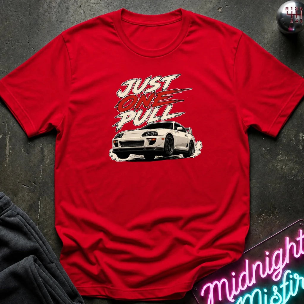 A red short-sleeve graphic T-shirt laid flat on a dark textured surface, featuring the words “JUST ONE PULL” in white and red above a white sports car illustration. 