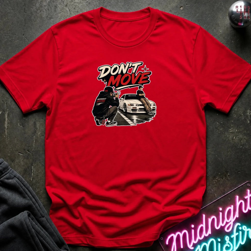A red short-sleeve graphic t-shirt laid flat on a dark textured surface. The graphic on the chest features the words 'DON'T MOVE' in a stylized white and red font above an illustration of a person and a white car.
