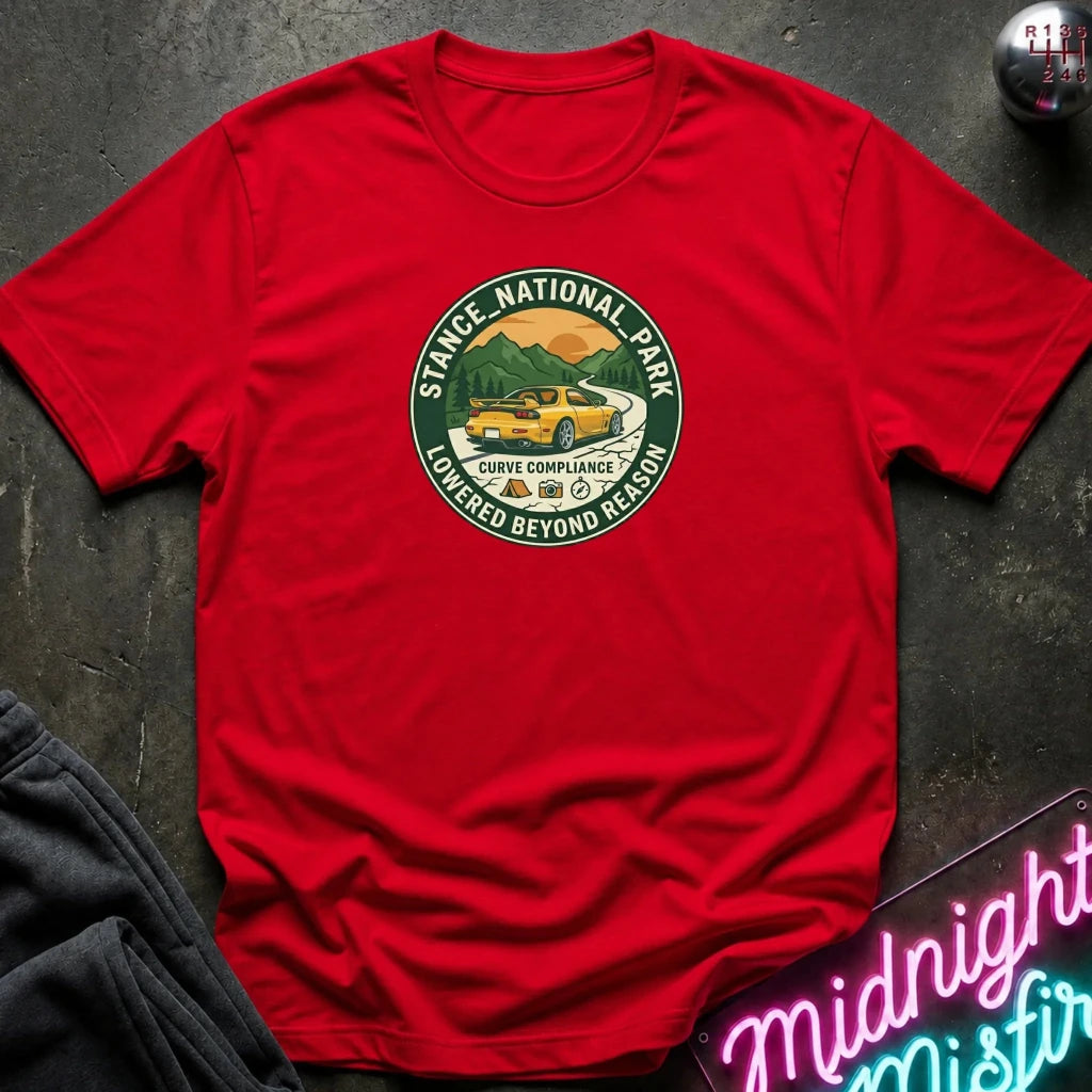 A red short-sleeve t-shirt laid flat on a dark textured surface, featuring a circular graphic print of a yellow car in a scenic mountain landscape with the text 'STACE NATIONAL PARK' and 'LOWEDGED BEYOND REASON'.