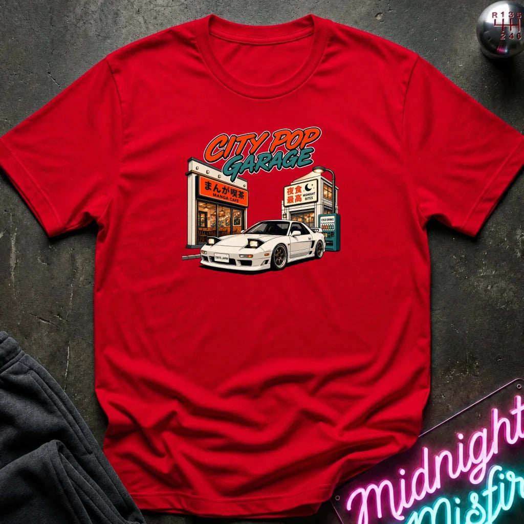A red short-sleeve graphic t-shirt laid flat on a dark textured surface. The graphic on the front shows a white sports car parked in front of a garage with stylized orange, yellow, and blue text reading 'CITY ROP GARAGE'.