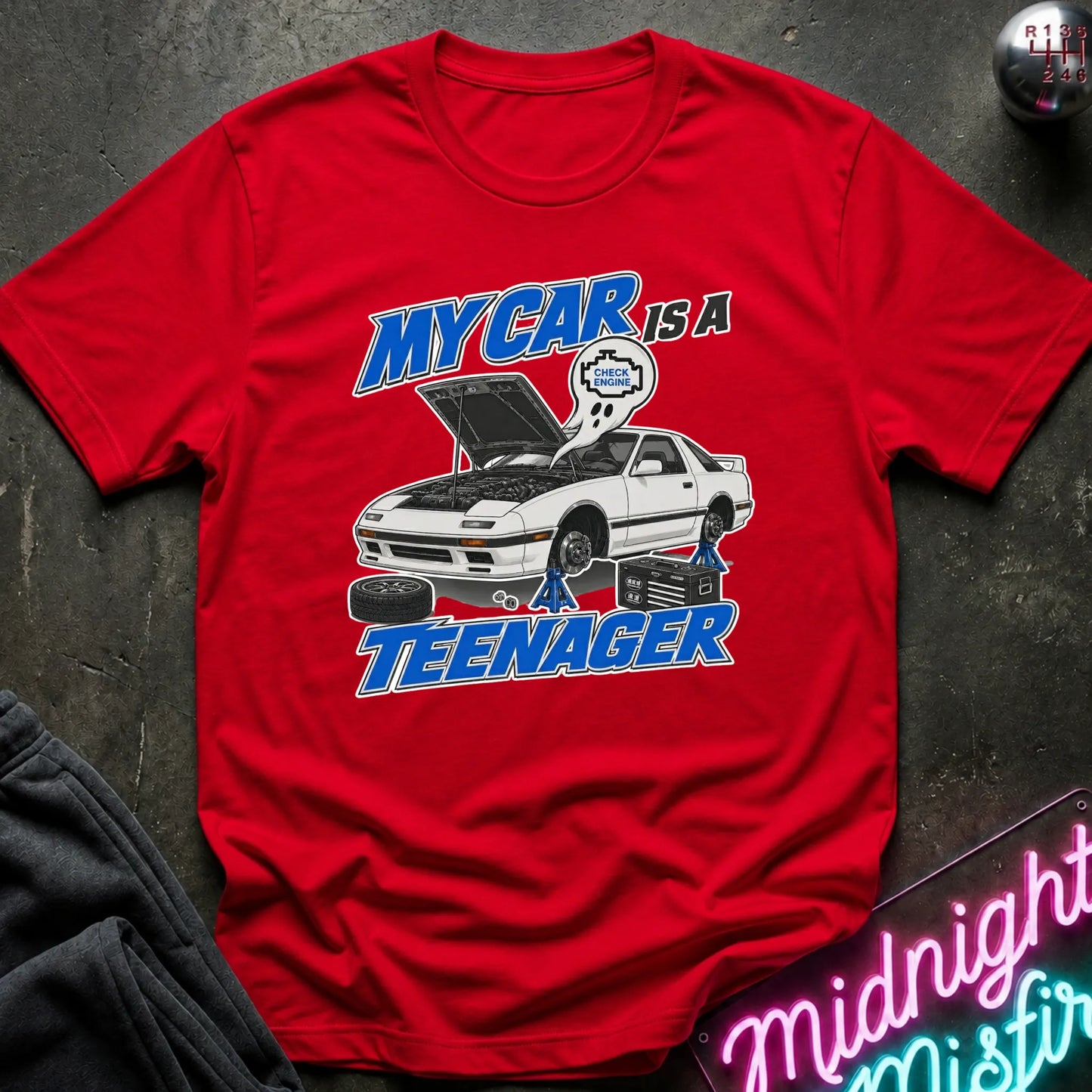 A red short-sleeve graphic T-shirt laid flat on a dark textured surface. The shirt features a white car with its hood open, blue and white text reading 'MY CAR IS A TEENAGER,'.