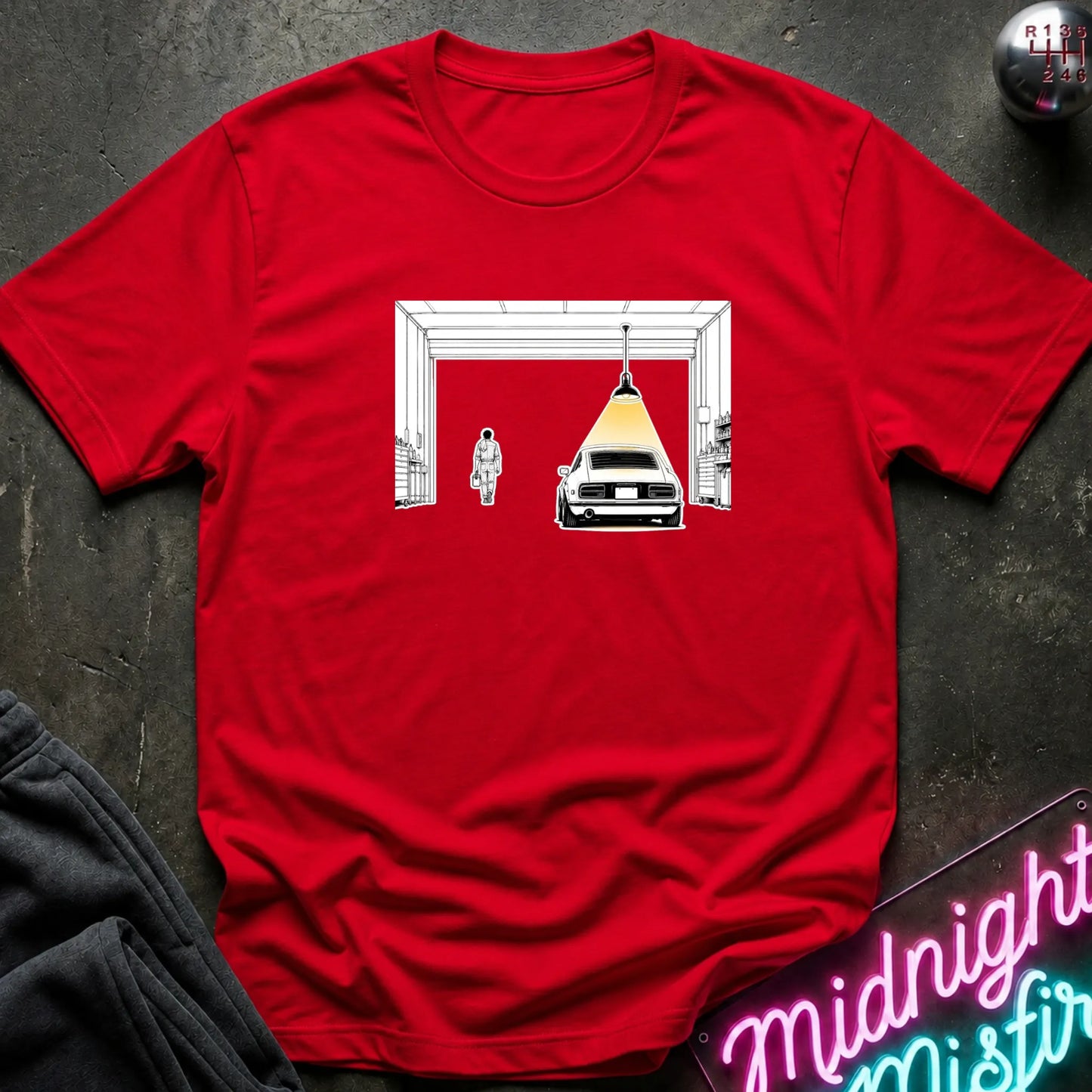 A red short-sleeve t-shirt laid flat on a dark textured surface, featuring a graphic print of a car under a hanging lamp inside a garage structure. 