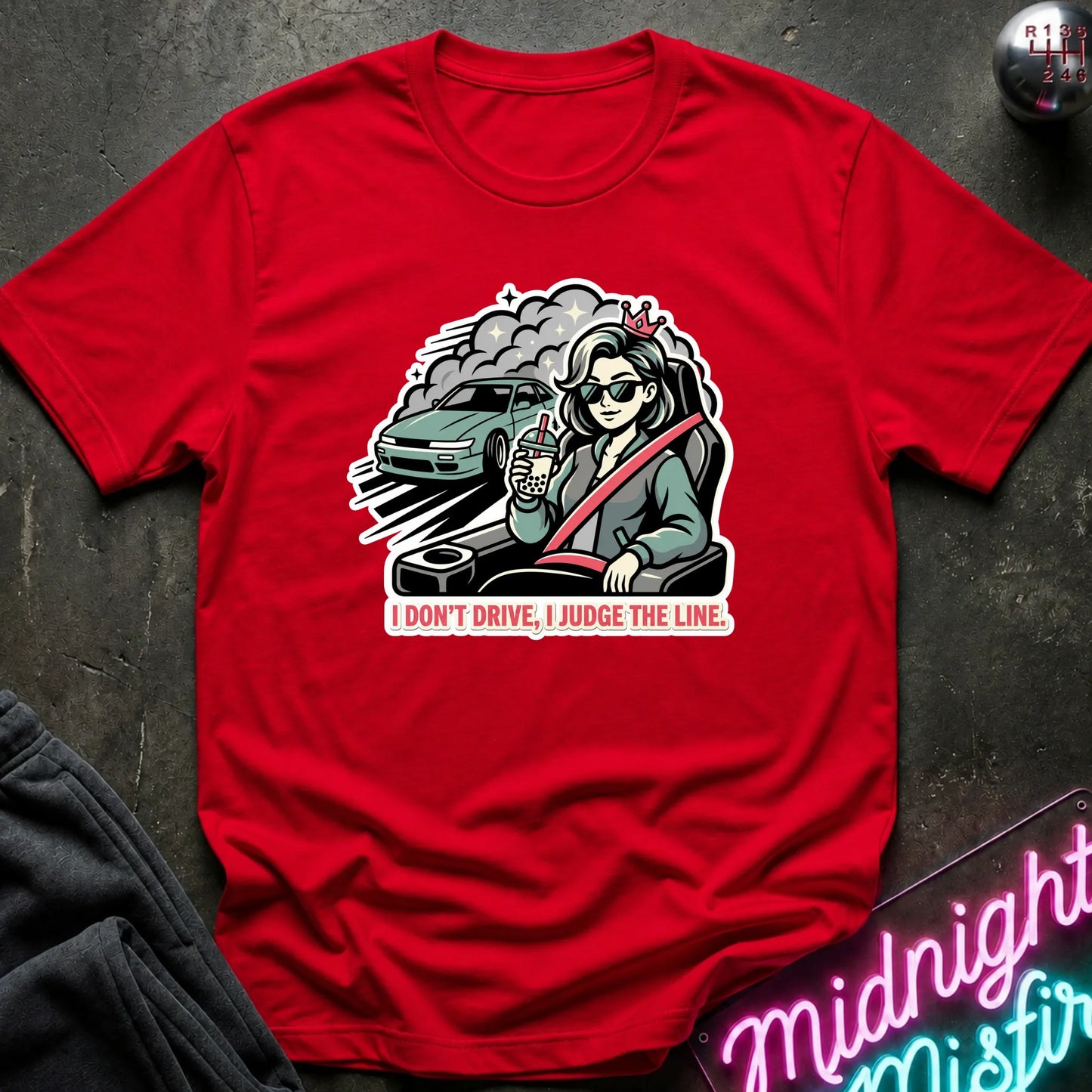 A red short-sleeve graphic t-shirt laid flat on a dark textured surface. The graphic shows a woman wearing sunglasses and a seatbelt with a car behind her and the text 'I DON'T DRIVE, I JUDGE THE LINE'.