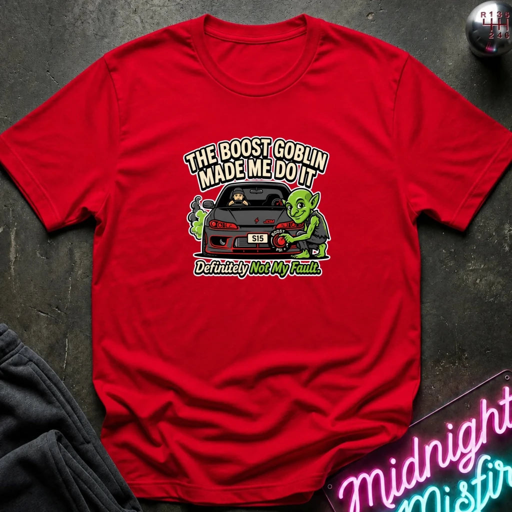 A red short-sleeve graphic t-shirt laid flat on a dark textured surface. The graphic features a green cartoon goblin next to a black car with the text 'THE BOOST GOBLIN MADE ME DOIT' and 'Definitely Not My Fault'.