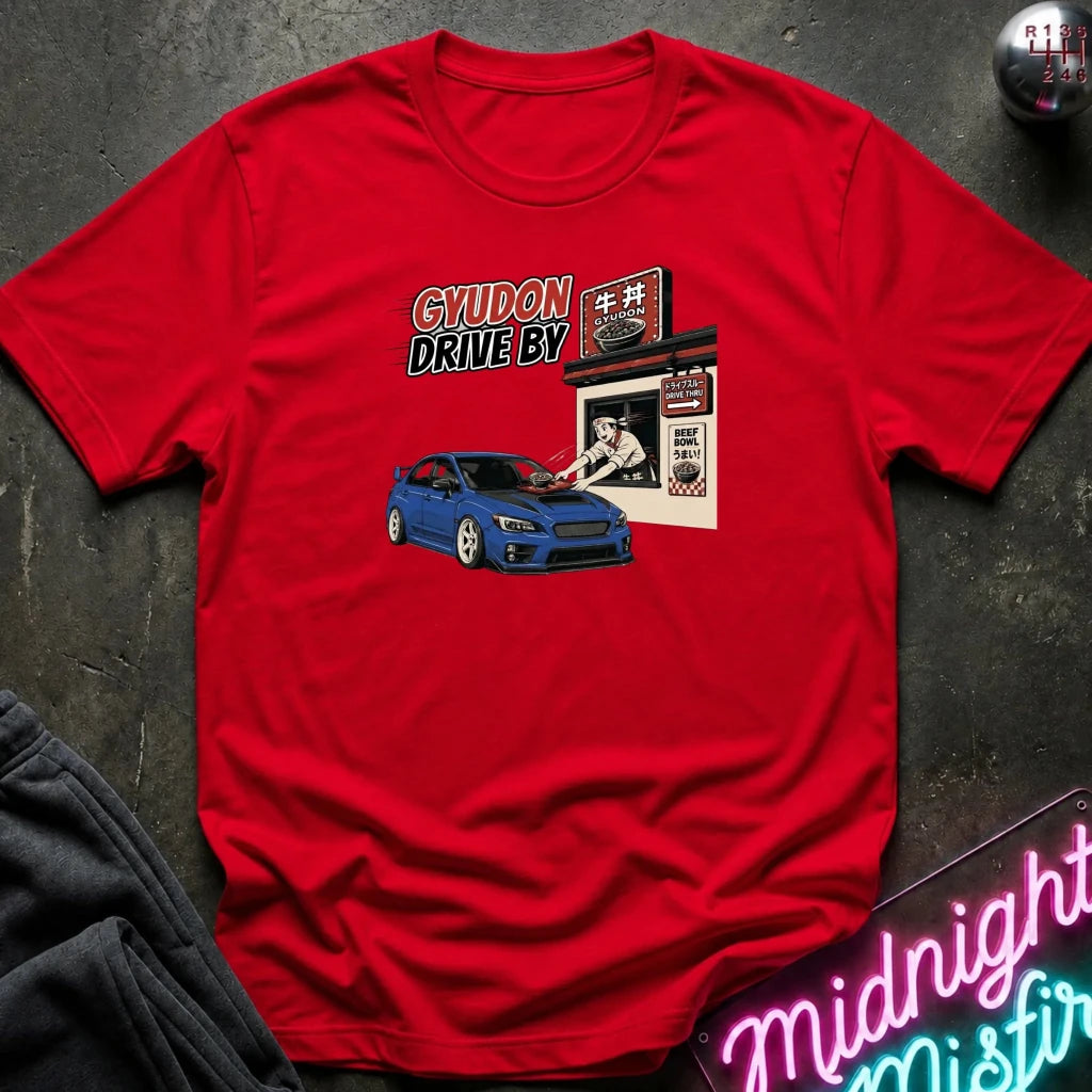 A red short-sleeve graphic t-shirt laid flat on a dark textured surface. The graphic features a blue sports car in front of a Japanese-style shop with the text 'GYUDON DRIVE BY'.