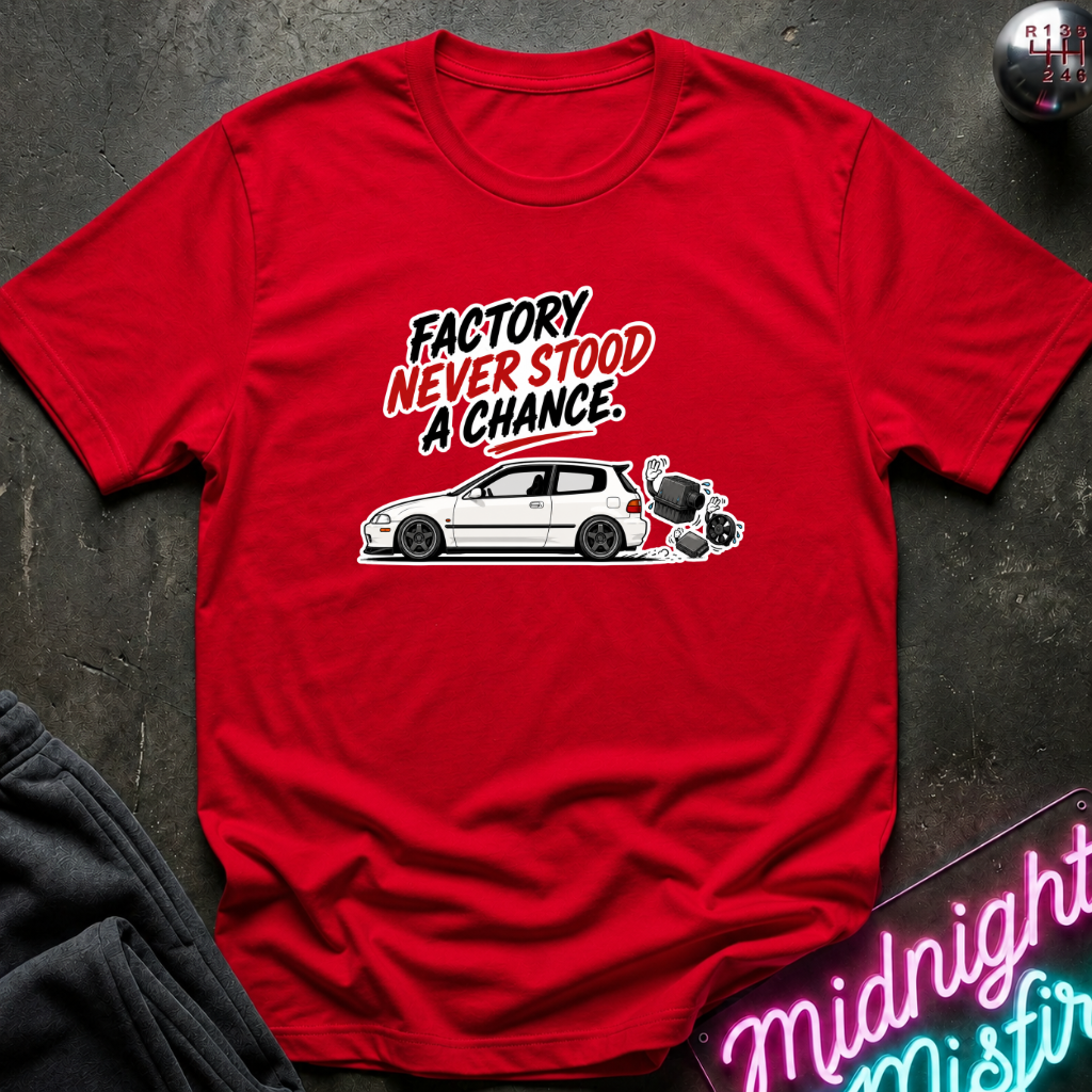 A red short-sleeve graphic T-shirt laid flat on a dark textured surface. The shirt features a white car illustration and the text 'FACTORY NEVER STOOD A CHANCE' in black and red lettering.