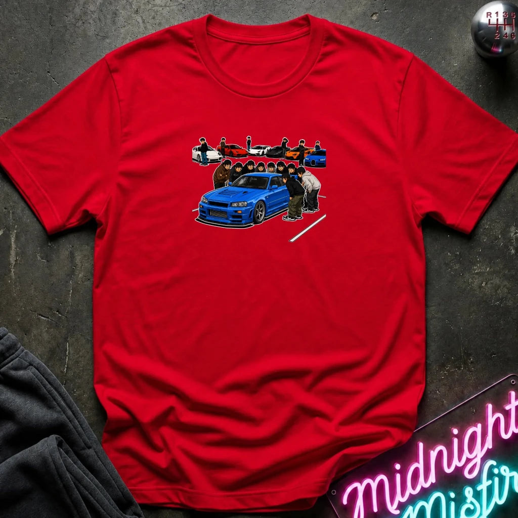 A red short-sleeve crewneck t-shirt laid flat on a dark textured surface. The center of the shirt features a graphic print of a blue sports car with a group of people standing behind it.