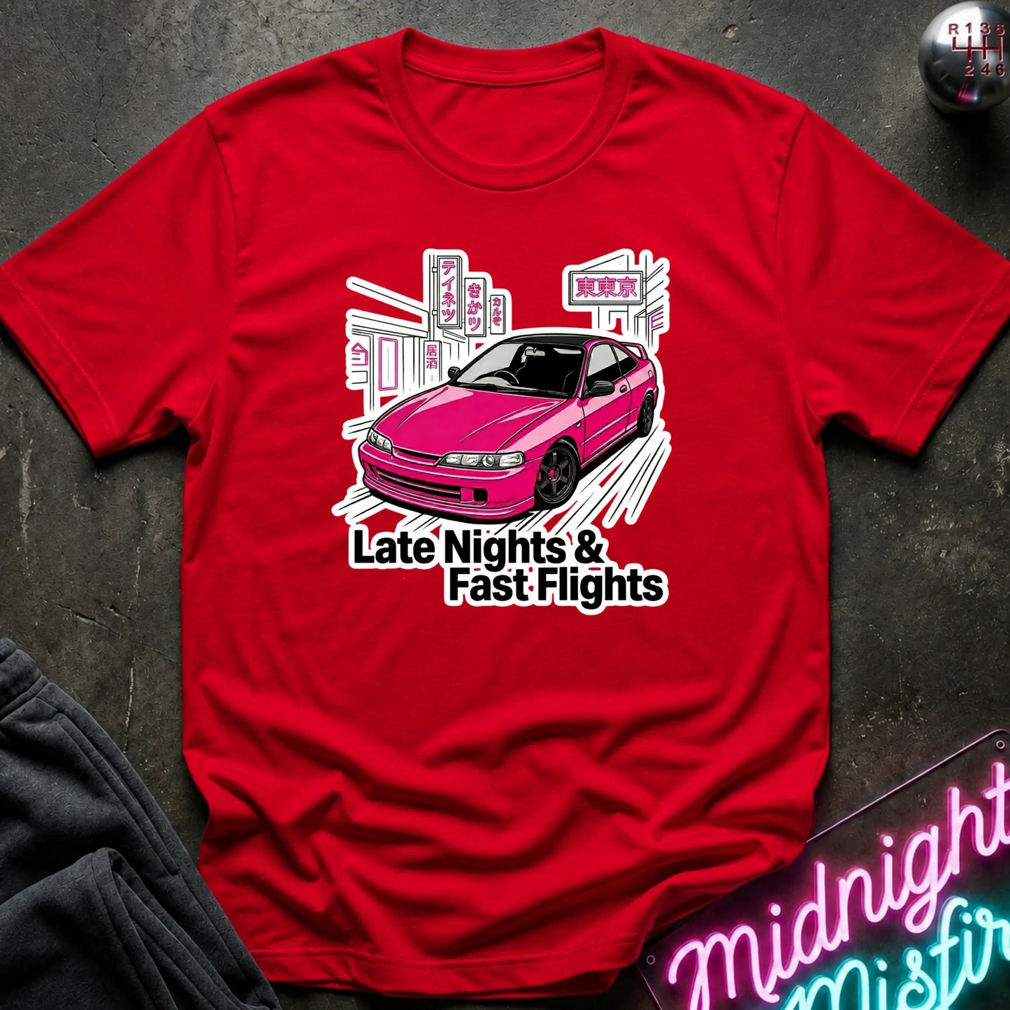  A red short-sleeve t-shirt laid flat on a dark textured surface, featuring a graphic of a pink car with the text 'Late Nights & Fast Flights'.