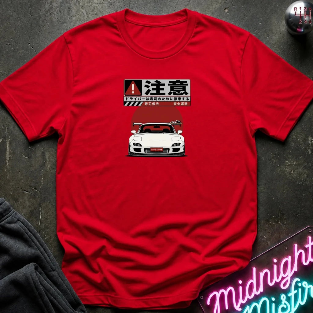 A bright red short-sleeve t-shirt laid flat on a dark textured surface. The shirt features a graphic print of a white sports car with Japanese text and a warning sign icon above it.
