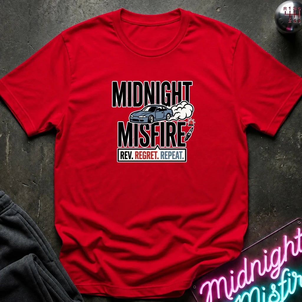 A red short-sleeve graphic T-shirt laid flat on a dark textured surface. The shirt features a central graphic with the words 'MIDNIGHT MISFIRE' in large black letters, a blue sports car, smoke, and a handbrake, with 'REV.