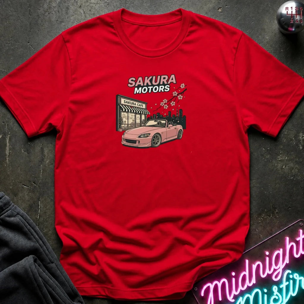 A red short-sleeve crewneck t-shirt laid flat on a dark textured surface. The shirt features a graphic print of a pink convertible car in front of a 'Sakura Motors' shop, with cherry blossoms and the text 'SAKURA MOTORS' above it.