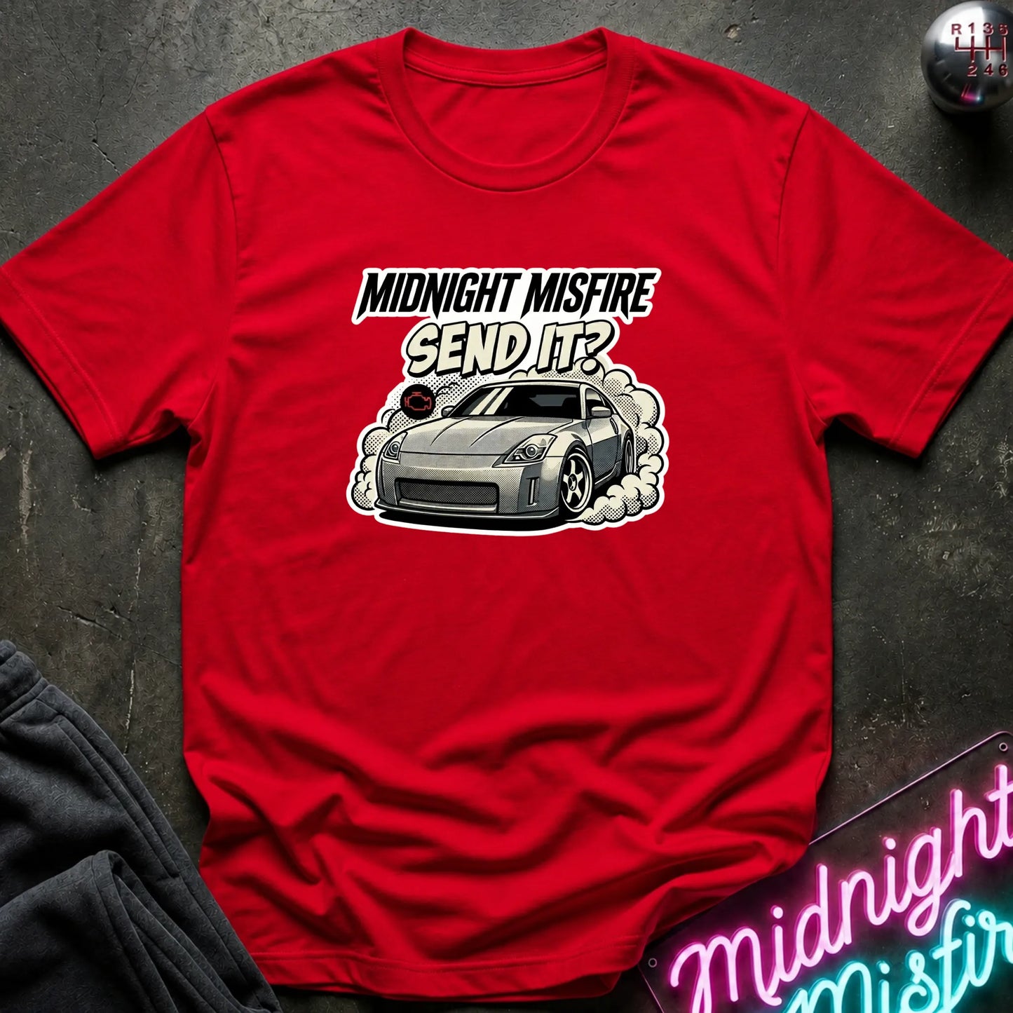 A red short-sleeve graphic T-shirt laid flat on a dark textured surface. The shirt features a printed design of a gray sports car with smoke and the text 'MIDNIGHT MISFIRE SEND IT?'.