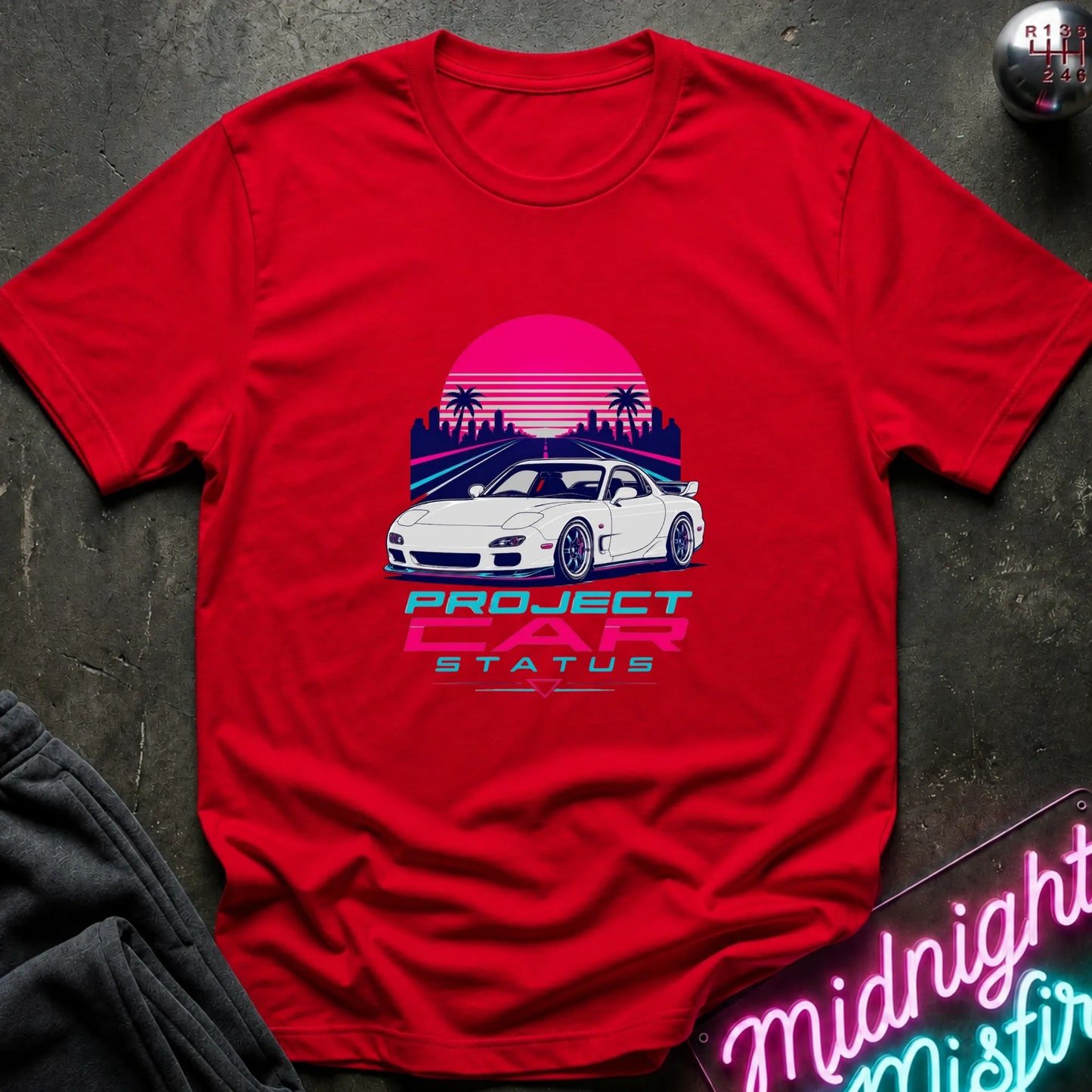 A red short-sleeve graphic T-shirt laid flat on a dark textured surface. The shirt features a retro-style print of a white sports car in front of a pink and purple sunset with palm trees, and the text 'PROJECT CAR STATUS' below.