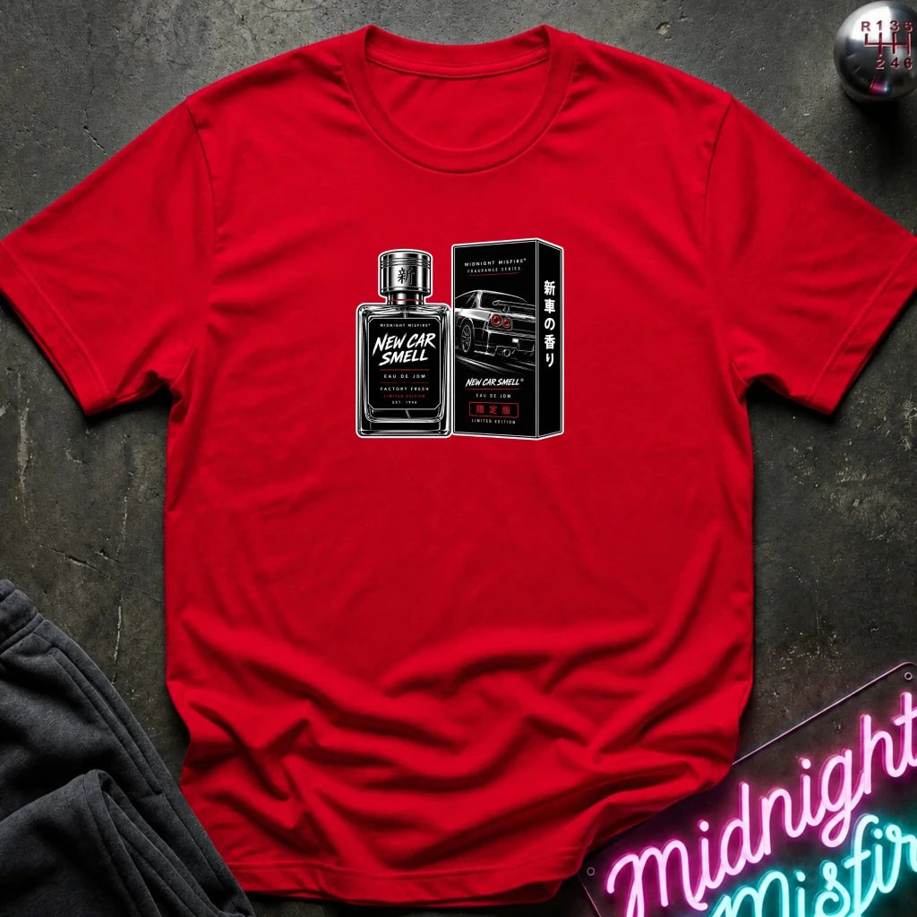 A red short-sleeve crewneck t-shirt laid flat on a dark textured surface, featuring a graphic print of a perfume bottle and box labeled 'New Car Smell' with a sports car. Decorative props include a neon sign in the corner and a dark sweatshirt edge.