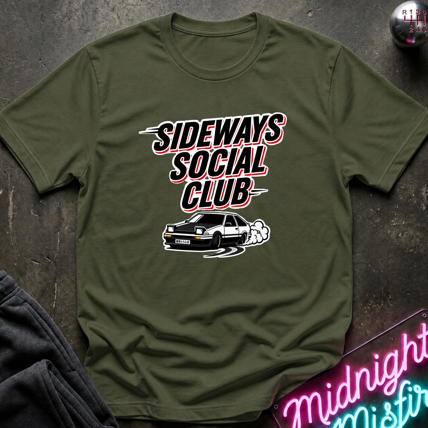 An olive green short-sleeve t-shirt with a graphic print that reads 'SIDEWAYS SOCIAL CLUB' in stylized black, white, and red lettering above a black car with white smoke. The shirt is laid flat on a dark textured surface with a neon sign and dark pants partially visible as props.