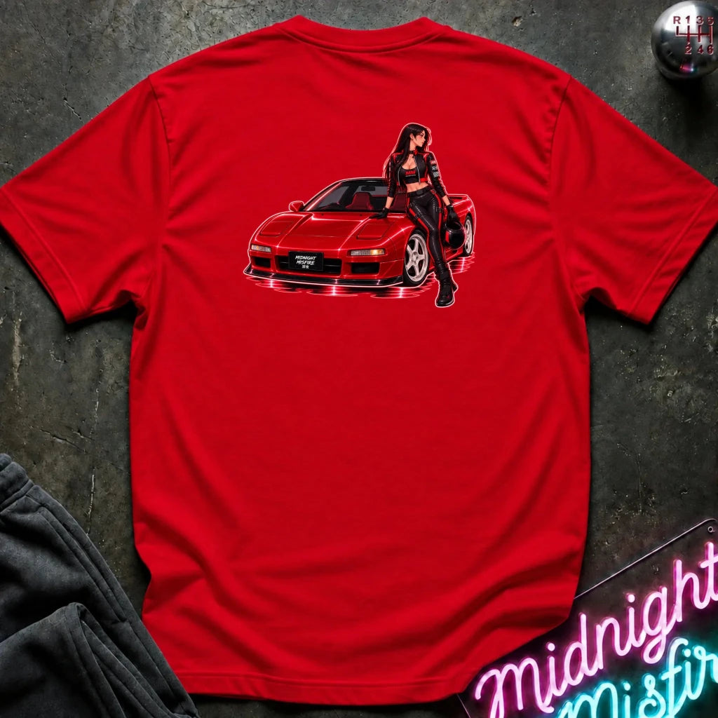 A red short-sleeve t-shirt laid flat on a dark textured surface, featuring a graphic print of a red sports car with a woman leaning on it on the back. A gear shift knob and a neon sign are partially visible as props.