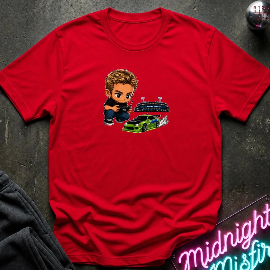 A red graphic t-shirt laid flat on a dark textured surface. The graphic features a green car, and person  controlling it with remote.
