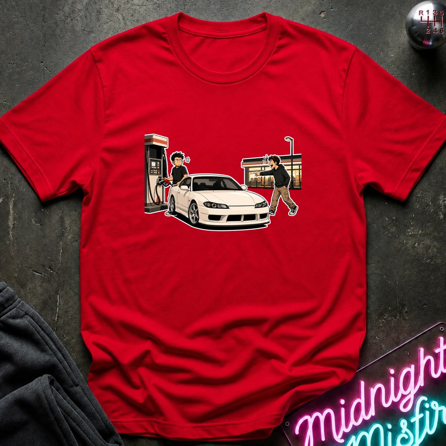 A red short-sleeve crewneck t-shirt featuring a cartoon graphic of a white sports car at a gas station, laid flat on a dark textured surface with a neon sign and dark sweatpants partially visible.