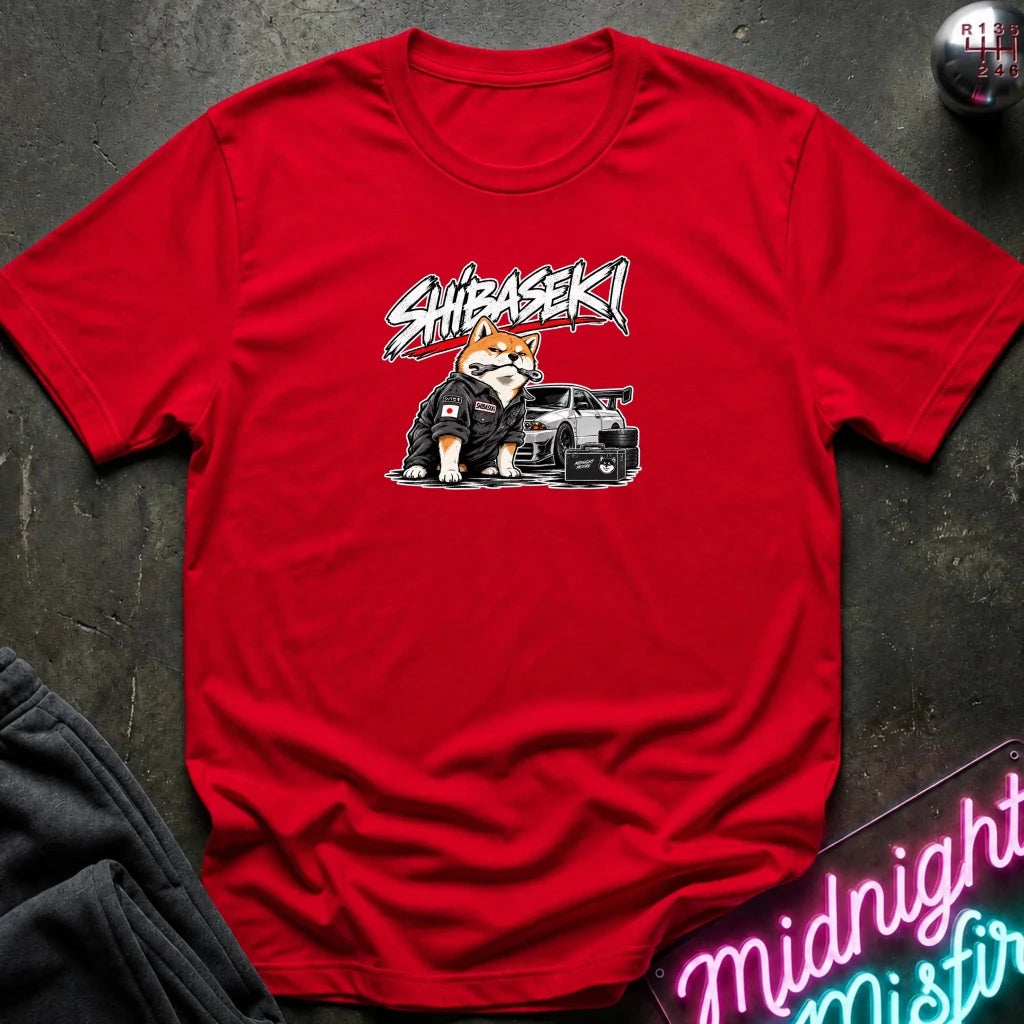 A red short-sleeve crewneck t-shirt laid flat on a dark textured surface. The center of the shirt features a graphic of a Shiba Inu dog wearing a black racing suit next to a black sports car, with the word 'SHIBASEKI' written above in a stylized white and black font.