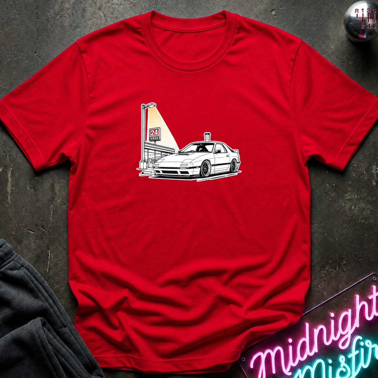 A red short-sleeve crewneck t-shirt laid flat on a dark textured surface, featuring a black line art graphic of a white sports car parked next to a lit street lamp.