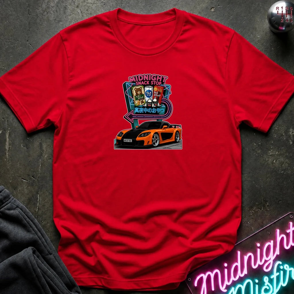 A red  short-sleeve t-shirt laid flat on a dark textured surface, featuring a colorful graphic print of an orange sports car in front of a neon sign that reads 'Midnight Snack Stop'.